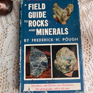 May include: A vintage field guide titled "A Field Guide to Rocks and Minerals" by Frederick H. Pough. The book has a blue cover with white text and illustrations of rocks and minerals. The cover also states it is the 3rd edition with 72 new color illustrations.