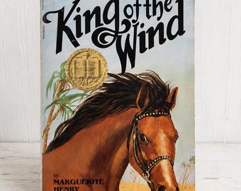 May include: A hardcover book titled King of the Wind by Marguerite Henry. The cover features a brown horse with a black mane wearing a bridle, set against a desert landscape with palm trees. A gold Newbery Medal seal is printed on the cover.
