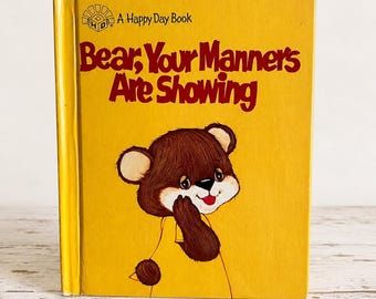 May include: A yellow children's book titled Bear, Your Manners Are Showing, featuring a Happy Day Book logo at the top and a brown bear illustration with a red tongue and dark brown fur centered on the front cover in a vertical orientation.