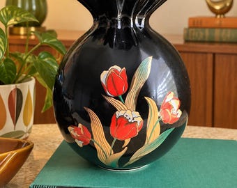 May include: A round, black ceramic vase with a ruffled, gold-rimmed opening. The front features a painted floral design of red tulips with green and gold leaves. The vase is resting on a green book titled The World of Washington Irving.