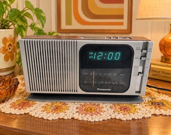 May include: A rectangular Panasonic AM/FM clock radio with a silver front panel, a dark wood-grain top, and a black display screen showing the time 12:00 in bright green digital numbers. The device features a vertical speaker grille and side controls.