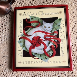 May include: A Cat's Christmas book by Stefanie Samek. The cover features a white cat wrapped in red ribbon on a green background with holly leaves. The book has a red border and the title is in a decorative font.