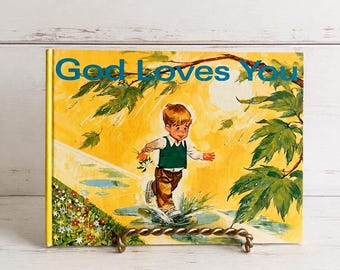 May include: A hardcover children's book titled God Loves You with a yellow cover illustration of a child in a green vest and brown pants jumping into a puddle. The scene includes green leaves, a bright sun, and colorful flowers in the corner.