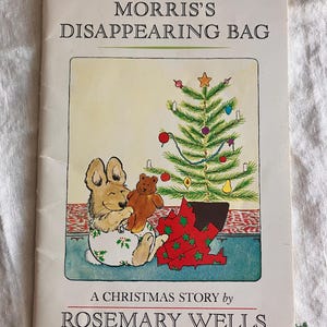 May include: A children's book cover titled "Morris's Disappearing Bag" by Rosemary Wells. The illustration features a rabbit holding a teddy bear near a Christmas tree and a wrapped present. The cover has a vintage, storybook aesthetic.