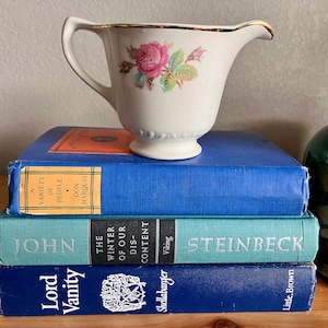 May include: A stack of three blue hardcover books with white text titles. The top book is titled "A Variety of People" by Don Marquis. The middle book is titled "The Winter of Our Discontent" by John Steinbeck. The bottom book is titled "Lord Vanity" by Shellabarger. A white creamer with a gold rim and a floral design sits on top of the books.