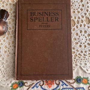 May include: A vintage brown book titled "BUSINESS SPELLER" with the author's name, Peters, visible. The book is set against a backdrop of white lace and embroidered fabric, suggesting a classic or antique theme.