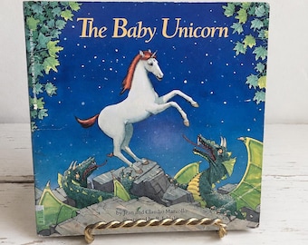 May include: A hardcover children's book titled The Baby Unicorn by Jean and Claudio Marzollo. The cover features a white unicorn with a red mane rearing on a stone structure, surrounded by two green dragons against a dark blue starry night sky.
