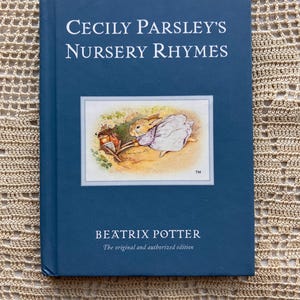 May include: A blue hardcover book titled Cecily Parsley's Nursery Rhymes by Beatrix Potter. The cover features a central illustration of a rabbit in a white garment pushing a small wooden cart, with the text The original and authorized edition.