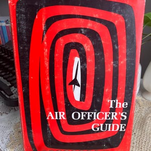 May include: Vintage book titled "The Air Officer's Guide" with a red and black spiral design on the cover. The book is from the 1965-1966 edition. The book is displayed on a gold-colored metal stand.