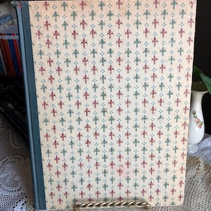 May include: A vintage book with a patterned cover featuring red, green, and blue fleur-de-lis designs on a cream background. The spine is a muted teal color. The book is displayed on a gold-toned metal stand.