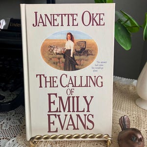 May include: A hardcover book titled "The Calling of Emily Evans" by Janette Oke. The cover features an illustration of a woman with a horse-drawn carriage. The book is displayed on a gold-colored metal stand.
