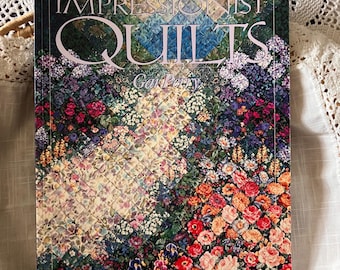 May include: A book titled "Impressionist Quilts" by Gai Perry. The cover features a colorful floral quilt design with red, pink, purple, and white flowers. The book is resting on a white, lace-like surface.