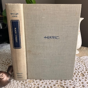 May include: A vintage copy of Sinclair Lewis's "Main Street" book. The book has a light beige spine with navy blue text and a light gray cover. The cover has the initials "HBMC" in navy blue. The book is part of the "Harcourt Modern Classics" series.