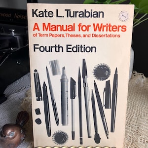 May include: A vintage copy of "A Manual for Writers" by Kate L. Turabian, Fourth Edition. The book cover is cream-colored with black and red text, and illustrations of pens and pencils. The book is displayed on a gold-toned stand.