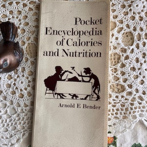 May include: A vintage paperback book titled "Pocket Encyclopedia of Calories and Nutrition" by Arnold E. Bender. The cover features a brown illustration of two figures at a table. The book is set against a white lace background.