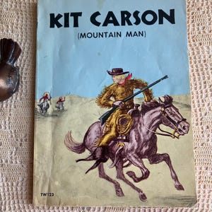 May include: A vintage softcover book titled KIT CARSON (MOUNTAIN MAN) with a light blue cover. The illustration depicts a person in fringed buckskin riding a brown horse while holding a rifle, with two figures on horseback in the background.