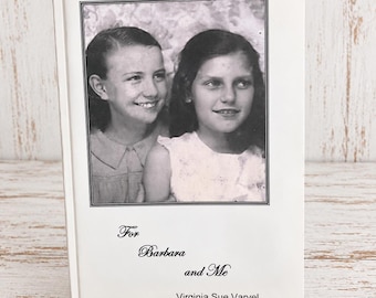 May include: A white hardcover book titled For Barbara and Me by Virginia Sue Varvel. The front cover features a black and white photograph of two young children smiling, framed by a thin black border. The book rests on a gold metal stand.
