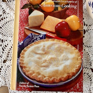 May include: A cookbook titled "The Illustrated Encyclopedia of American Cooking" featuring a pie, cheese, apples, and a knife. The cover has a red background with the title in white. The book is from the Editors of Favorite Recipes Press.