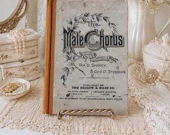 May include: A vintage book titled The Male Chorus, composed and arranged by Ira D. Sankey and Geo. C. Stebbins, published by The Biglow & Main Co. The cover is light gray with black text and a botanical illustration, featuring a brown spine.