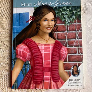 May include: A book titled Meet Marie-Grace 1853 from American Girl featuring an illustration of a girl with brown hair and blue eyes wearing a pink plaid dress with red ruffles. The cover includes a small inset photo and the text The Story Continues! See back cover.