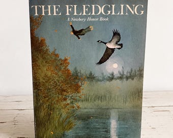 May include: A paperback book titled The Fledgling by Jane Langton, a Harper Trophy Book. The cover features an illustration of a person flying alongside a Canada goose over a lake at twilight, with autumn foliage on the left and a full moon.