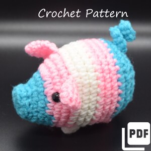 May include: Crochet pattern for a small pig amigurumi toy. The pig is crocheted in blue, pink, and white yarn, creating a transgender pride flag color scheme. The pattern is available as a PDF download.