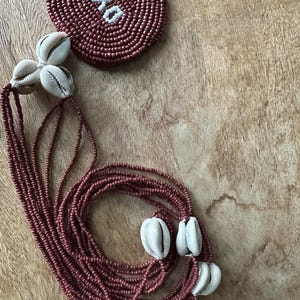 May include: A multi-strand necklace featuring dark red seed beads and white cowrie shells. A circular beaded medallion is attached to the strands, displaying the word OYA in white beads against a dark red background, creating a distinct contrast.