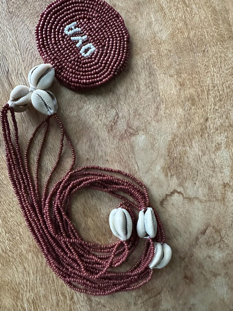 May include: A multi-strand necklace featuring dark red seed beads and white cowrie shells. A circular beaded medallion is attached to the strands, displaying the word OYA in white beads against a dark red background, creating a distinct contrast.