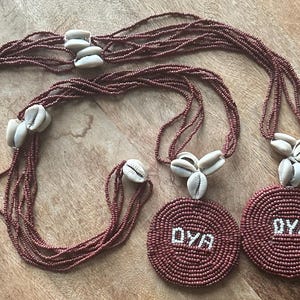 May include: Two multi-strand necklaces made of small, dark reddish-brown beads. Each necklace features a circular pendant with the white beaded text OYA, accented by clusters of off-white cowrie shells along the strands and above the pendants.