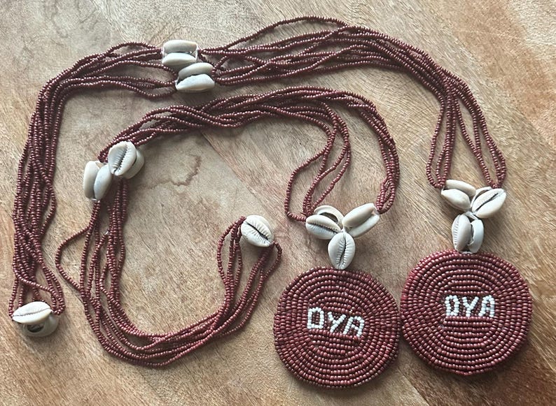 May include: Two multi-strand necklaces made of small, dark reddish-brown beads. Each necklace features a circular pendant with the white beaded text OYA, accented by clusters of off-white cowrie shells along the strands and above the pendants.