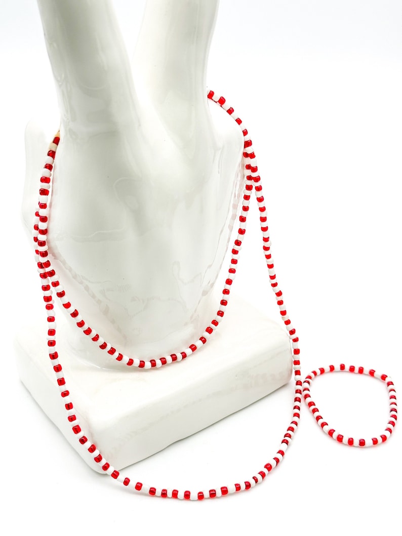Op de afbeelding: Een lange ketting en een bijpassende armband gemaakt van kleine, afwisselend rode en witte cilindrische kralen. De sieraden hebben een eenvoudig, uniform patroon van individuele rode en witte kralen die aan een dun, flexibel koord zijn geregen.
