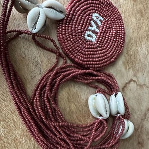 May include: A multi-strand necklace made of small, dark mauve beads featuring clusters of white cowrie shells. Attached is a circular, flat medallion crafted from the same mauve beads with the word OYA spelled out in white beads at the center.