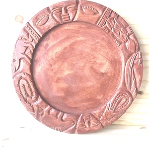 Peut inclure: Une assiette circulaire en bois brun avec un centre plat et lisse et un large bord surélevé. Le bord est orné de motifs en relief sculptés à la main, incluant des visages humains stylisés et diverses formes géométriques sur tout le pourtour.