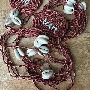 May include: A set of two circular beaded medallions and long strands of dark rose-colored seed beads accented with white cowrie shells. Each medallion features the word "OYO" stitched in white beads against a dark rose background.