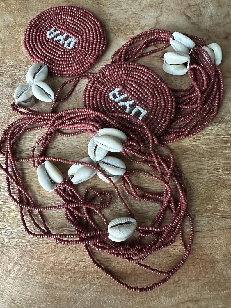May include: A set of two circular beaded medallions and long strands of dark rose-colored seed beads accented with white cowrie shells. Each medallion features the word "OYO" stitched in white beads against a dark rose background.