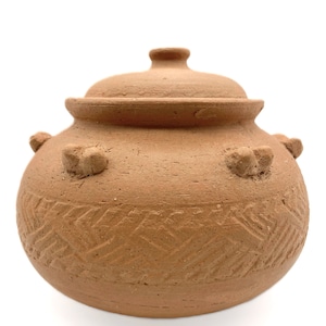 May include: A terracotta-colored ceramic pot with a lid. The pot has a rounded body with a textured, etched pattern around the base. It features three decorative handles and a knob on the lid. The pot is likely used for cooking or storage.