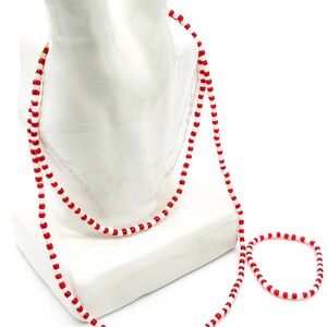 Op de afbeelding: Een lange ketting en een bijpassende armband gemaakt van kleine, afwisselend rode en witte cilindervormige kralen. De kralen zijn geregen aan een dun koord, waardoor een herhalend patroon van rood en wit ontstaat in beide sieraden.