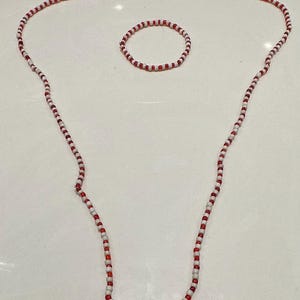 Op de afbeelding: Een lange ketting en een bijpassende ronde armband gemaakt van kleine, afwisselend rode en witte kralen. De kralen zijn uniform van grootte en vorm, waardoor een eenvoudig, herhalend patroon ontstaat in beide sieraden van deze set.