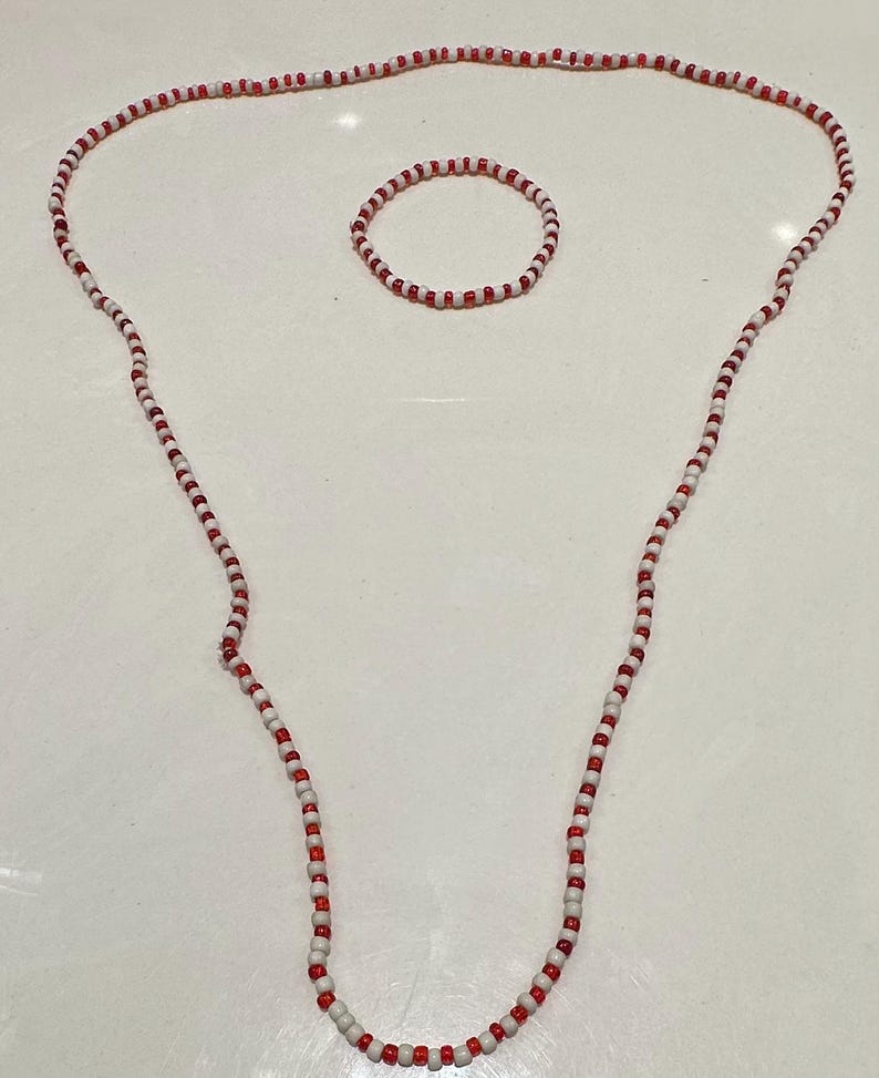 Op de afbeelding: Een lange ketting en een bijpassende ronde armband gemaakt van kleine, afwisselend rode en witte kralen. De kralen zijn uniform van grootte en vorm, waardoor een eenvoudig, herhalend patroon ontstaat in beide sieraden van deze set.