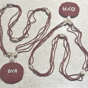 May include: A multi-strand necklace featuring deep red seed beads and off-white cowrie shells. The design includes two circular beaded medallions, each displaying the white text OYA, connected by long, layered strands of red beads and shell accents.