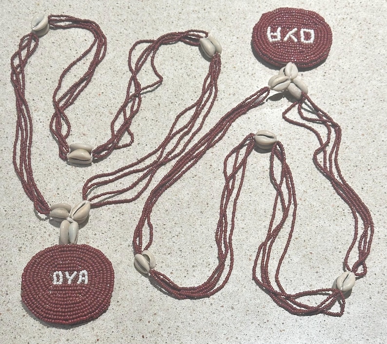 May include: A multi-strand necklace featuring deep red seed beads and off-white cowrie shells. The design includes two circular beaded medallions, each displaying the white text OYA, connected by long, layered strands of red beads and shell accents.