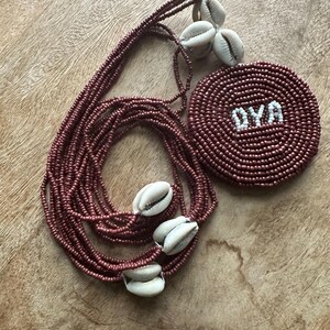 May include: A multi-strand necklace made of small, metallic maroon beads featuring several white cowrie shells and a circular, flat pendant. The pendant is crafted from the same maroon beads and displays the white text OYA centered on its surface.