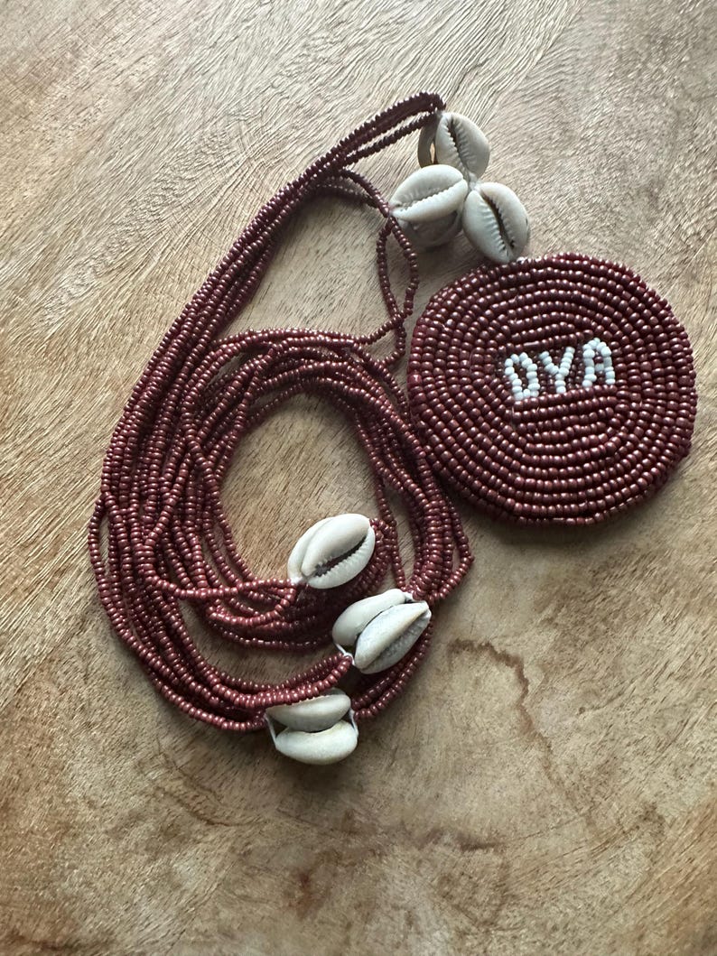 May include: A multi-strand necklace made of small, metallic maroon beads featuring several white cowrie shells and a circular, flat pendant. The pendant is crafted from the same maroon beads and displays the white text OYA centered on its surface.