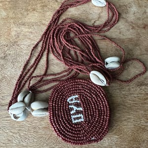 May include: A multi-strand necklace featuring small, dark reddish-brown beads and several white cowrie shells. A large, circular, dark reddish-brown beaded pendant is attached, displaying the word OYA in white beads at its center.