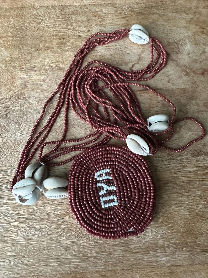 May include: A multi-strand necklace featuring small, dark reddish-brown beads and several white cowrie shells. A large, circular, dark reddish-brown beaded pendant is attached, displaying the word OYA in white beads at its center.