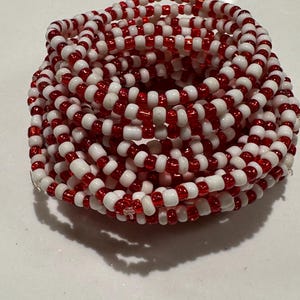 Op de afbeelding: Een opgerolde armband gemaakt van talloze kleine, ronde kralen in afwisselende rode en witte kleuren. De kralen zijn geregen in een doorlopend, meerlaags cirkelvormig patroon, wat zorgt voor een dicht, getextureerd uiterlijk met kleurcontrast.