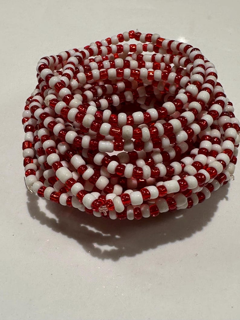 Op de afbeelding: Een opgerolde armband gemaakt van talloze kleine, ronde kralen in afwisselende rode en witte kleuren. De kralen zijn geregen in een doorlopend, meerlaags cirkelvormig patroon, wat zorgt voor een dicht, getextureerd uiterlijk met kleurcontrast.