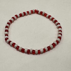 Op de afbeelding: Een cirkelvormige kralenarmband met een afwisselend patroon van kleine, ronde, doorschijnende rode kralen en ondoorzichtige witte kralen. De kralen zijn geregen in een doorlopende lus, wat zorgt voor een eenvoudig, uniform ontwerp met een herhalende kleurvolgorde.