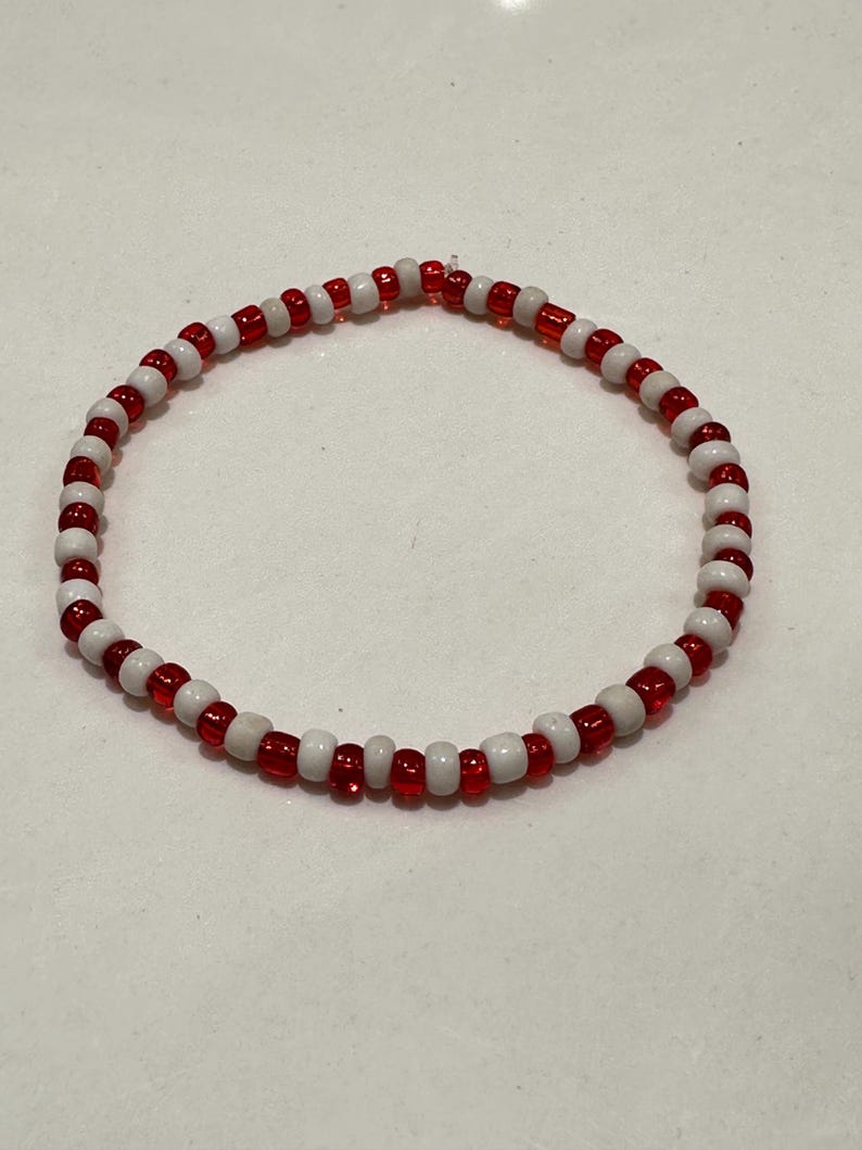 Op de afbeelding: Een cirkelvormige kralenarmband met een afwisselend patroon van kleine, ronde, doorschijnende rode kralen en ondoorzichtige witte kralen. De kralen zijn geregen in een doorlopende lus, wat zorgt voor een eenvoudig, uniform ontwerp met een herhalende kleurvolgorde.