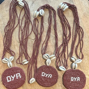 May include: Three necklaces featuring multiple strands of small, dark red beads. Each necklace has a circular pendant made of dark red beads with the white text OYA centered on it. The pendants are accented with three white cowrie shells.